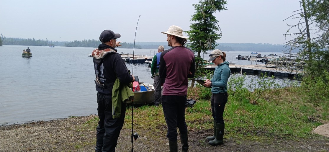 An employee of the Aquatic Invasive Species Awareness Team chats with a boater holding a fishing rod on the shore of a lake.
